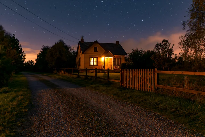 A wooden house stands along a gravel driveway under a night sky filled with stars.