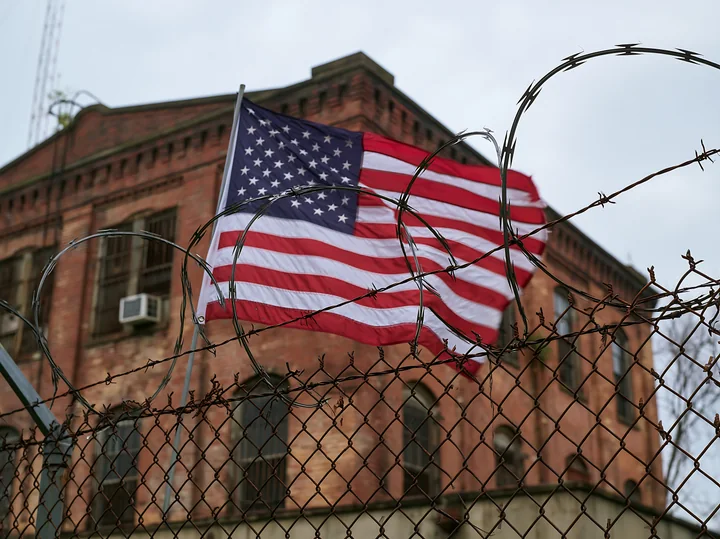 An American flag behind barbed wire is visible near an old brick building.