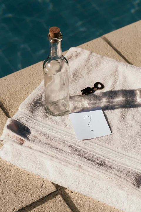 An empty bottle with question mark note sits on a white towel near a swimming pool.