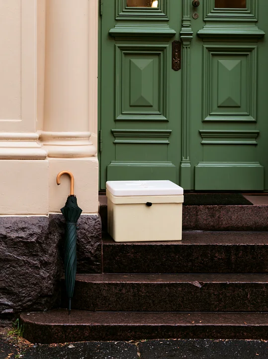 An insulated milk delivery box rests beside an umbrella on the steps of an apartment building with green doors.