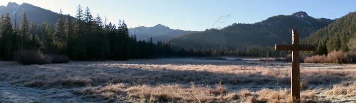 A carved oak cross stands in a frosted meadow surrounded by tall trees and mountains. The scene captures the early morning light and the stillness of nature in