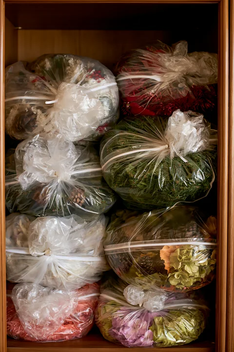 Clear bags of Christmas decorations, greenery, tinsel, pinecones, and flowers fill a wooden storage cabinet.
