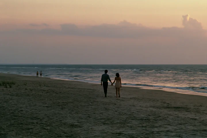 Couple holding hands while walking along a quiet beach beside the ocean at dusk.