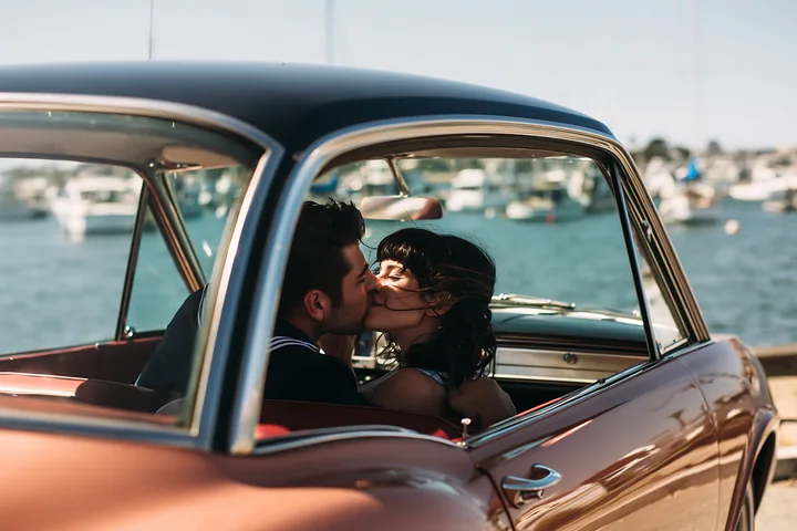 Couple kissing inside a vintage car beside a sunny waterfront.