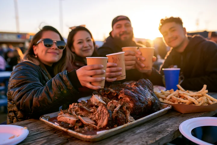 Friends celebrate at a pig roast at a food festival with plates of food and drinks in hand.