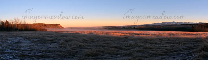Frosty meadow in a rural area during sunrise with soft light illuminating a misty landscape. Mountains in the distance create a scenic backdrop. This scene capt