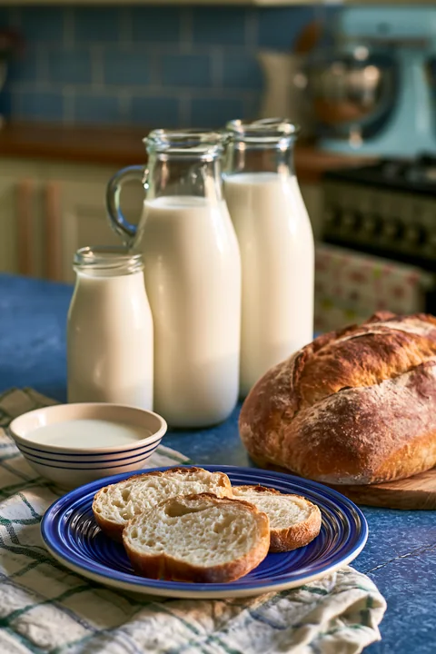 Glass milk bottles are lined up next to slices of fresh bread on a blue table.