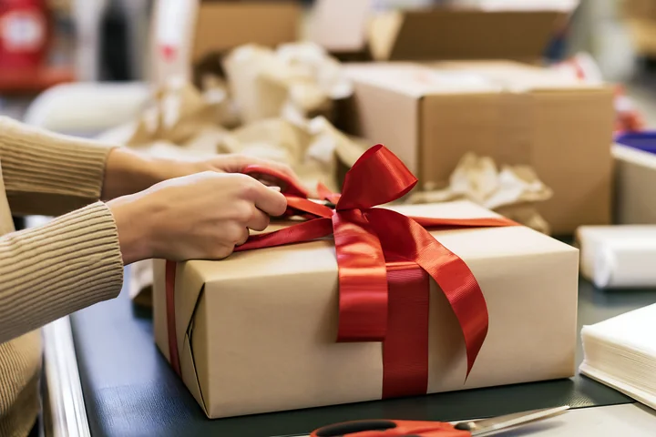 Hands tie a red ribbon around a wrapped gift box on a worktable.