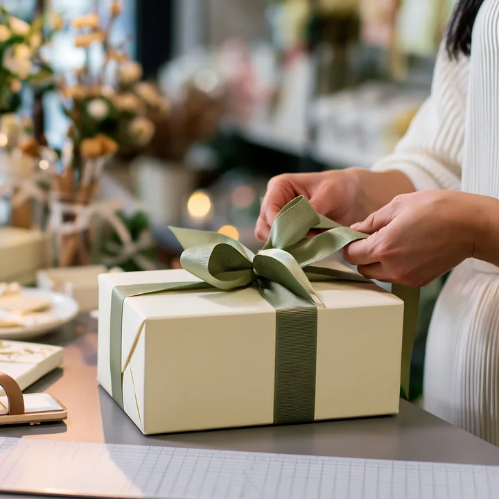 Hands tying ribbon around gift box in a shop filled with decorations and gift items.