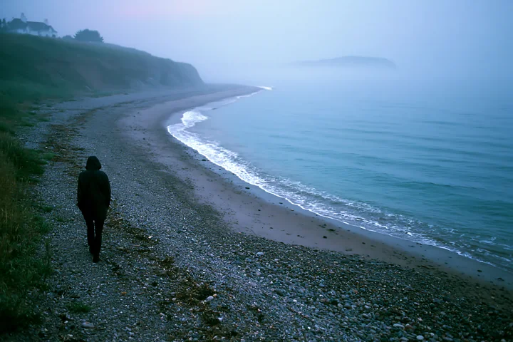 Hooded person walks alone along a misty pebble beach beside calm coastal water and cliffs.