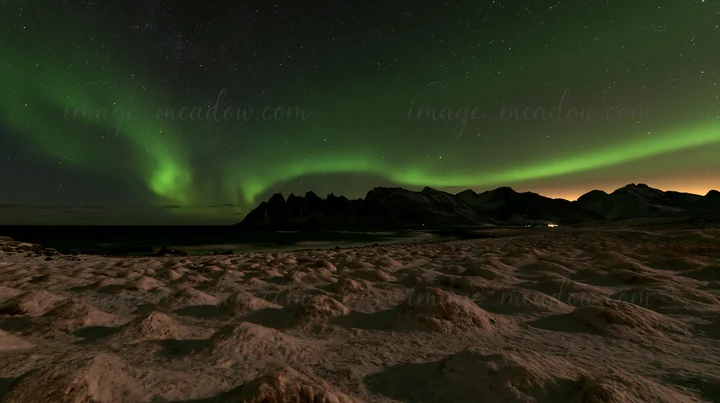 Scenes show a coastal meadow in Lofoten during winter, covered in snow. Northern lights illuminate the sky, creating a beautiful contrast against the dark mount