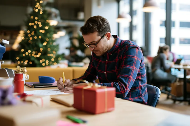Man writing a gift list at a desk beside wrapped presents and a decorated Christmas tree.