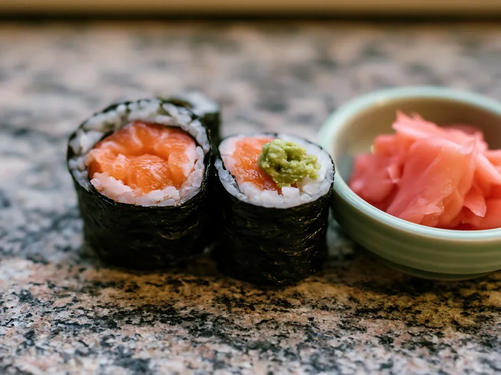 One salmon maki piece with wasabi sits on a granite countertop alongside a small bowl of pickled ginger.