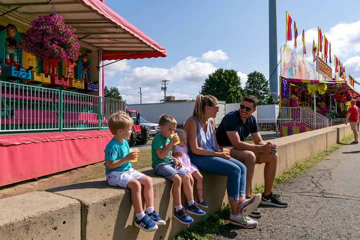 Parents and three children sit beside colorful fairground game stalls while enjoying drinks on a sunny day.
