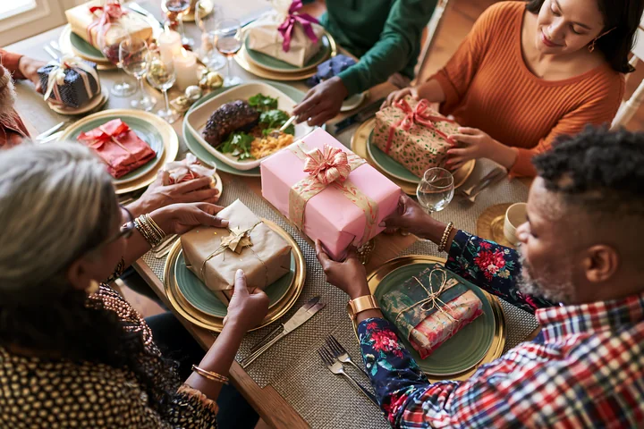 Parents arrange wrapped gifts around a dining table set for a festive family meal.