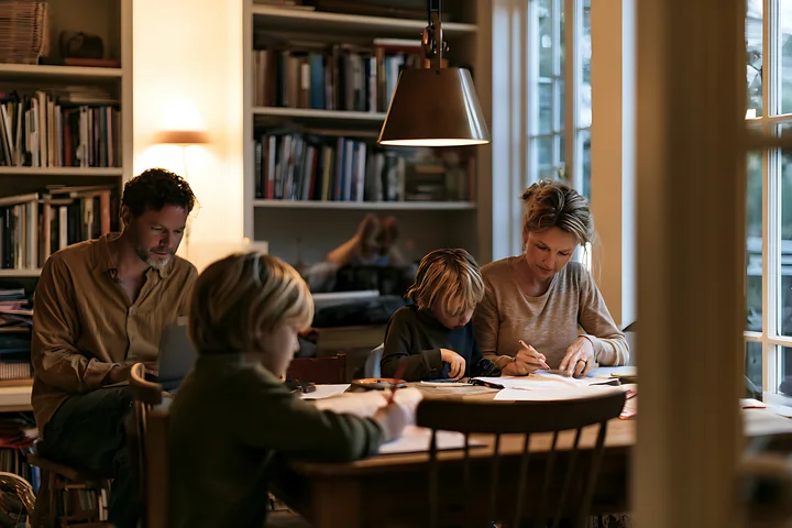 Parents work at home while two children study at a dining table surrounded by bookshelves.