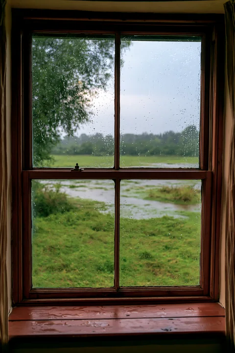 Rain on small cottage window overlooks a green field with water pooling in the grass.