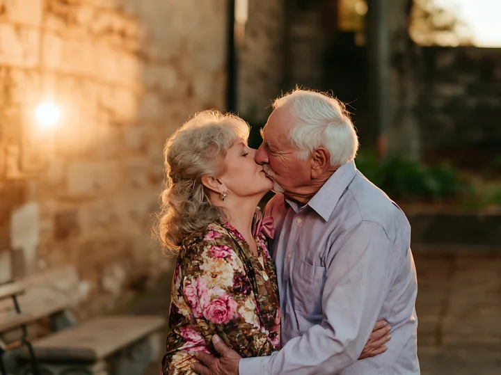 Senior couple kissing outdoors at sunset, warmly lit beside a stone wall with open copy space to the left.