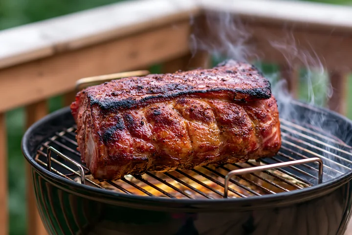 Smoke rises from a barbecue grill where a whole roast pork is cooking on an outdoor deck.