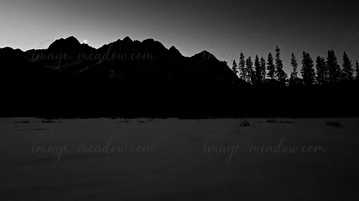 A snowy meadow in Rocky Mountain Park is seen during dusk. Tall trees frame the area, with mountains rising in the background under a twilight sky. Presented as