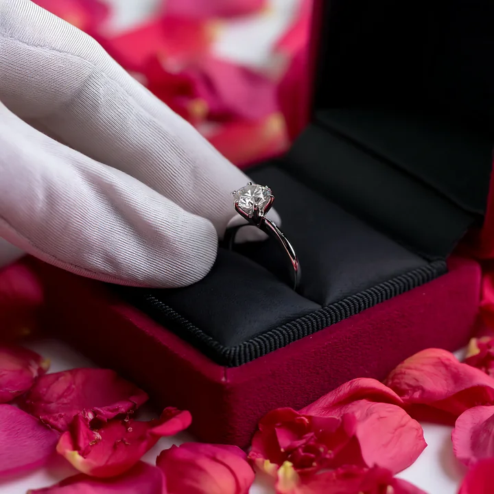 Sparkling engagement ring in an open black jewelry box beside scattered pink flower petals and a white glove.