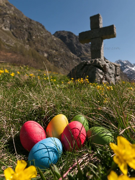 A stone cross stands in an alpine valley meadow surrounded by flowers. Colorful eggs in pink, blue, yellow, and green lie on the grass, indicating a festive atm
