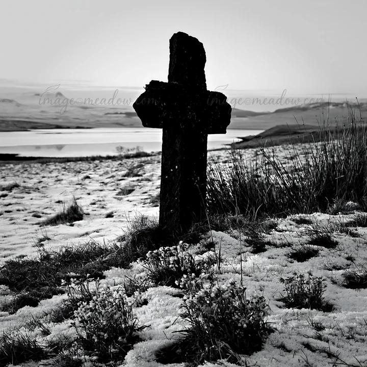 A stone Christian cross stands in a snowy meadow with patches of grass. The landscape features a quiet body of water in the background and distant hills. The se