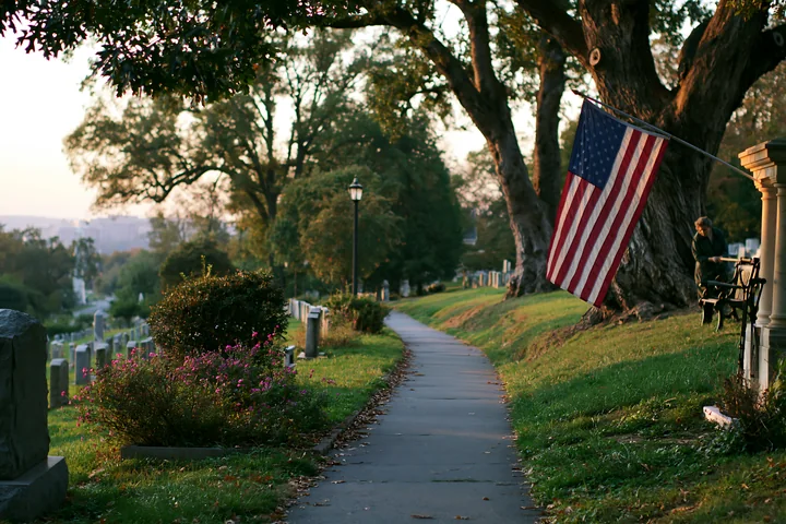 The American flag at Arlington Cemetery is seen along a quiet path lined with gravestones and trees during the golden hour of sunset.