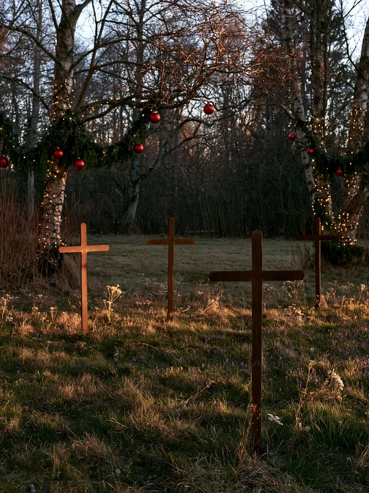 Three iron crosses stand in an Advent meadow. The setting includes trees decorated with lights and ornaments. The scene captures a quiet moment in the late afte