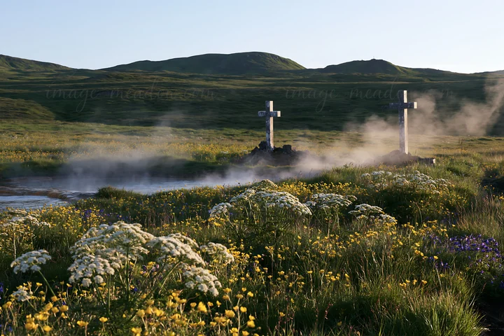 Two Christian crosses stand in a meadow with blooming flowers near a thermal spring. Steam rises from the spring, and hills are visible in the background during