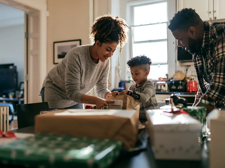 Two parents and their child wrap gifts together at a dining table in a bright home kitchen.