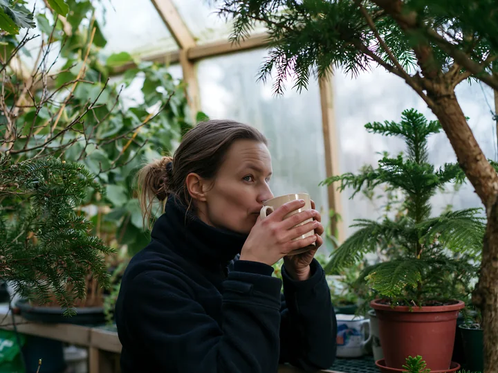 Woman drinks cocoa from a mug while surrounded by plants inside a bright greenhouse.