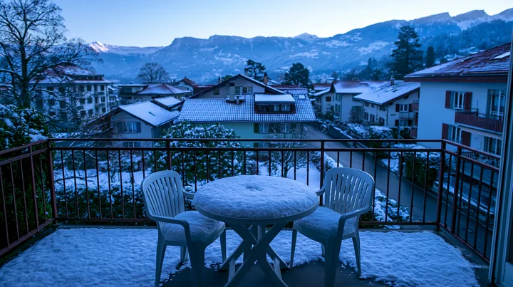 A balcony with two chairs and a round table covered in snow offers a view of Swiss mountain scenery with houses and trees in winter.