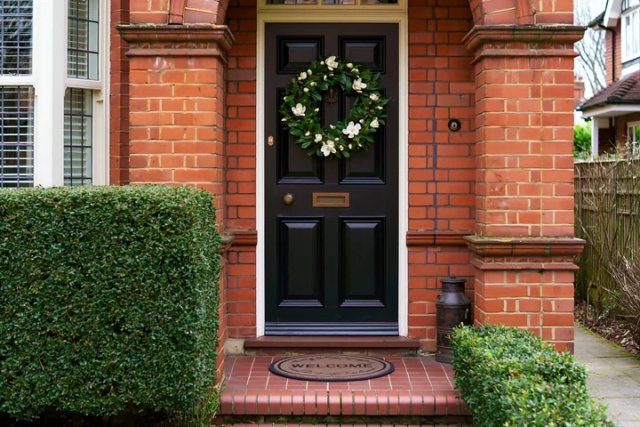 A black front door features a green wreath and a welcome doormat at a brick home entrance.