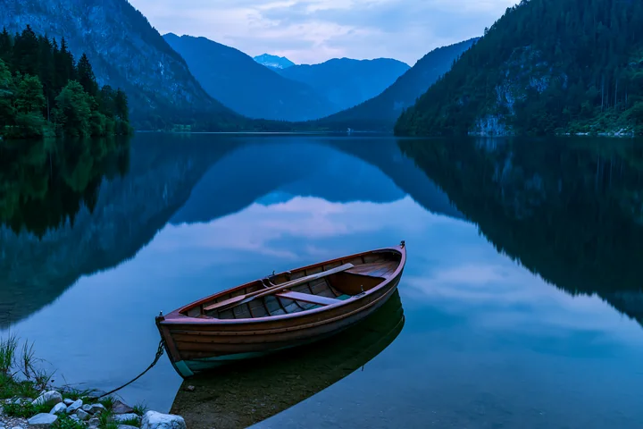 A boat is moored at the edge of an alpine lake, which reflects the surrounding mountains and trees.