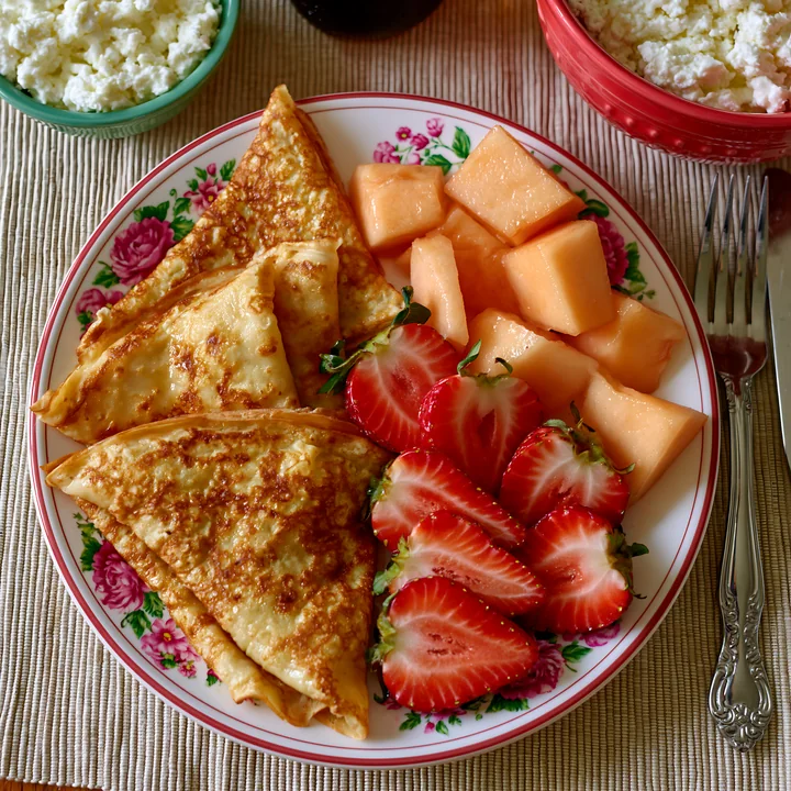 A breakfast plate features sweet crepes alongside fresh strawberries and melon cubes.