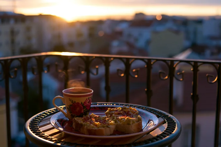 A breakfast plate with avocado toast and a cup of coffee sits on a balcony table as the sun rises over the city skyline.