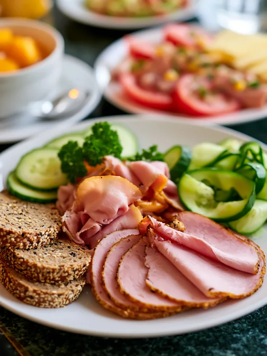 A breakfast plate with cold cuts includes slices of ham, turkey, and bread alongside cucumber, parsley, and a side of yogurt.
