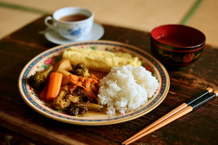 A breakfast plate with Japanese breakfast includes rice, vegetables, and an omelet alongside a cup of tea, arranged on a wooden table in a traditional setting.