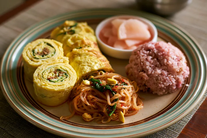 A breakfast plate with Korean breakfast includes rolled omelets, stir-fried noodles, pink rice, and pickled radish.