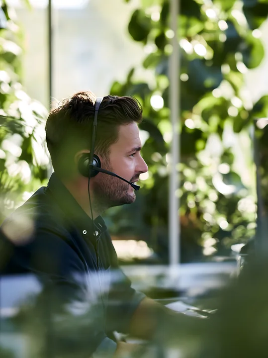 A businessman wearing a headset talks on a phone in an office filled with plants.