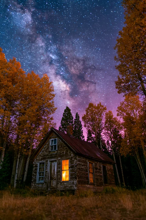 A cabin stands in a valley with bright stars shining above and trees in autumn colors surrounding it.