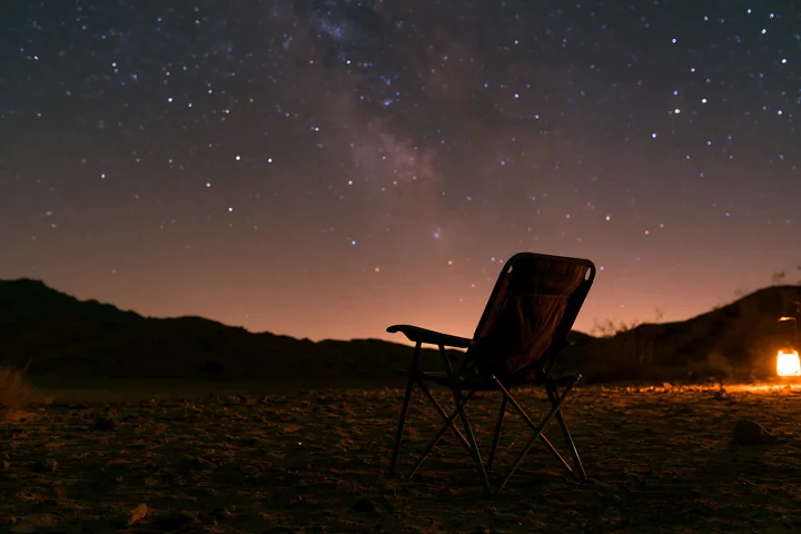 A camping chair sits on the ground under a starry night above a desert campsite, with a lantern providing light nearby and dark mountains in the background.