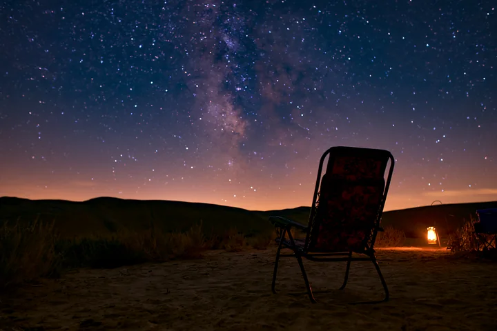 A chair sits on the sand of a desert campsite under a starry night sky.