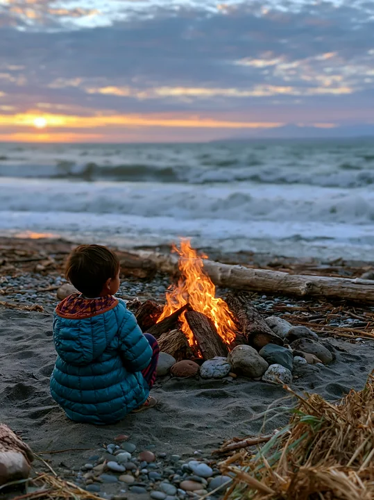 A child sits by a beach bonfire, watching the flames as waves crash on the shore at sunset.