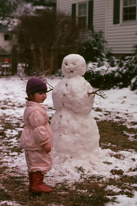 A child stands beside a snowman in a snowy backyard.