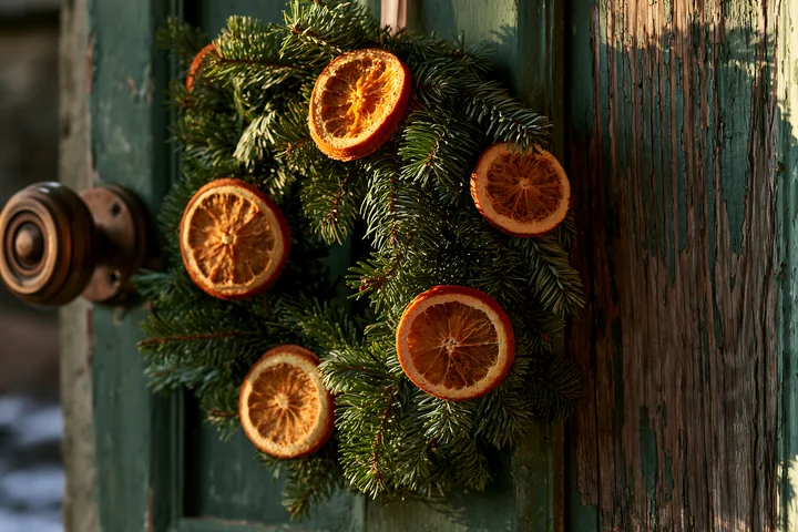 A Christmas wreath made of evergreen branches and dried orange slices hangs on a wooden apartment door, adding a festive touch to the entrance.