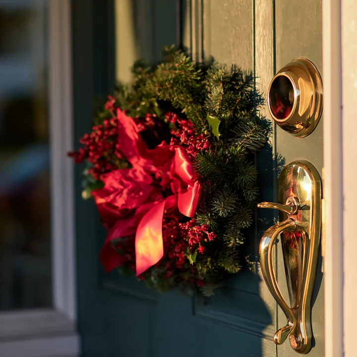A Christmas wreath with red decorations hangs on a green apartment door beside a shiny gold handle.