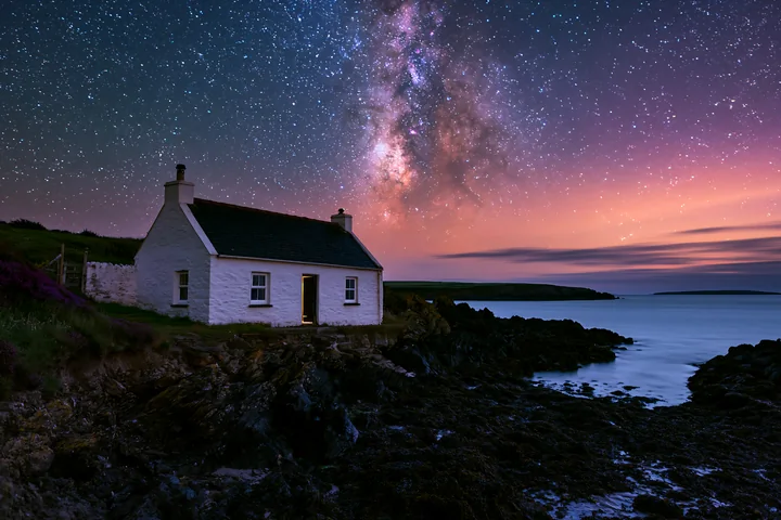 A coastal cottage stands near the rocky shore as stars fill the sky.