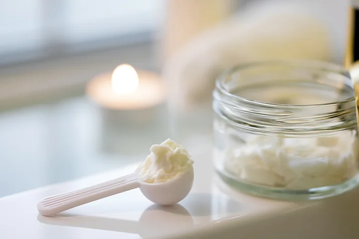 A cosmetic scoop holds a cream dose next to a glass jar of cream on a bathroom counter.